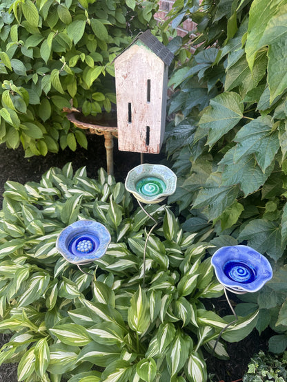 Three JJ Potts Studio ceramic Flower Puddlers in blue and green staked among hostas beside a wooden birdhouse
